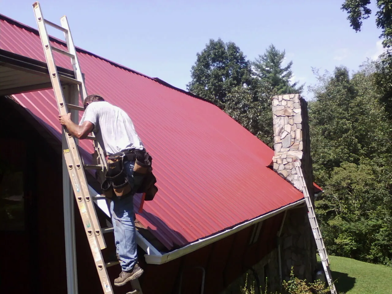 Roofer on a ladder beside a red metal roof and stone chimney — Soffit & Fascia Repair by Roofing Brothers in Nickerson